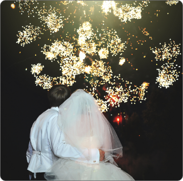 A bride and groom sitting in front of fireworks.